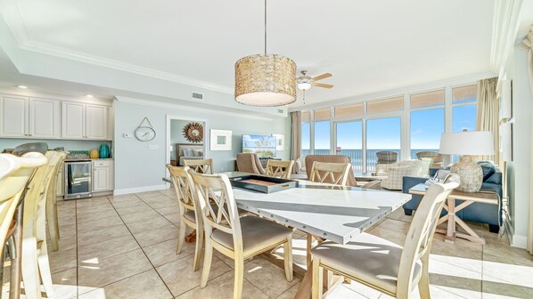 A spacious living area with a large dining table, comfortable seating, a ceiling fan, and a view of the ocean through large windows.