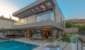 Ein modernes, zweistöckiges Haus mit Glasfassade, Sitzgelegenheit im Freien und einem Swimmingpool.