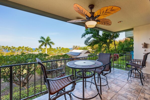 A balcony with a table and chairs, a ceiling fan, and a view of palm trees and the ocean.