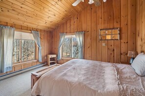 A wooden cabin bedroom with a large bed, two windows with curtains, a small wooden bench, and a wall-mounted light fixture.