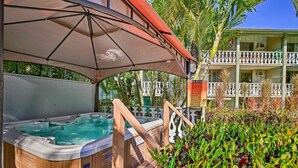 A spa area with a hot tub under a canopy, surrounded by lush greenery and a building with balconies. - Nautical Theme Garden View Studio - Kona Islander Inn Condos Condo by RedAwning (Kailua-Kona)