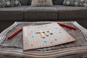A Scrabble board on a woven tray with tiles and a red rack, set on a wicker table in front of a grey sofa with patterned cushions.