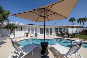 A swimming pool with lounge chairs and an umbrella, surrounded by a house and palm trees.