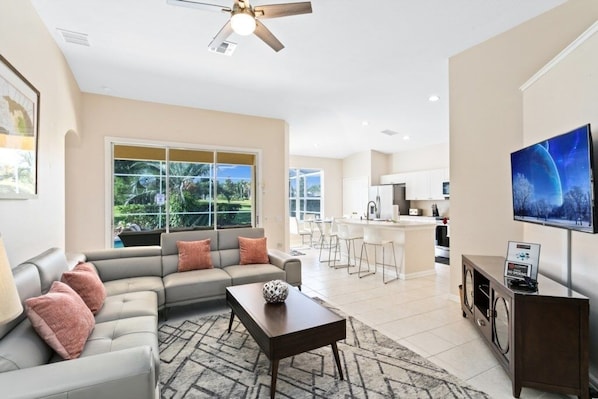 A modern living room with a sofa, coffee table, and a TV. There is a kitchen area in the background with a dining set.