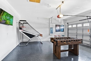 A room with a foosball table, a ping pong table, and a wall-mounted TV.