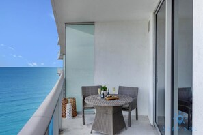 A balcony with a round wicker table, two chairs, and a glass door leading to a pool. - Oceanfront Elegance: Sunset Views, Pet-Friendly (Hollywood)