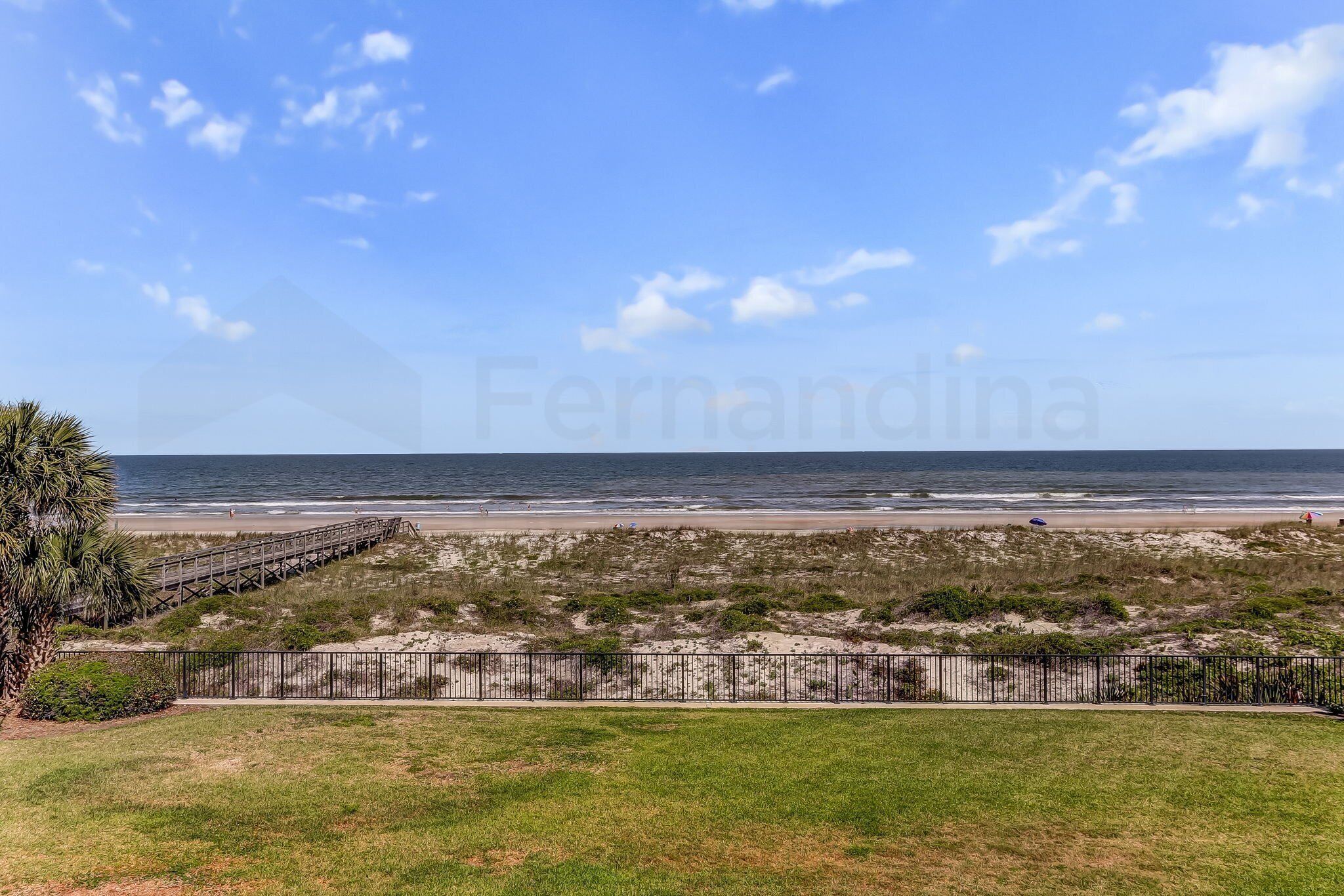 Vista de una playa con un paseo marítimo de madera, zonas verdes y una cerca.