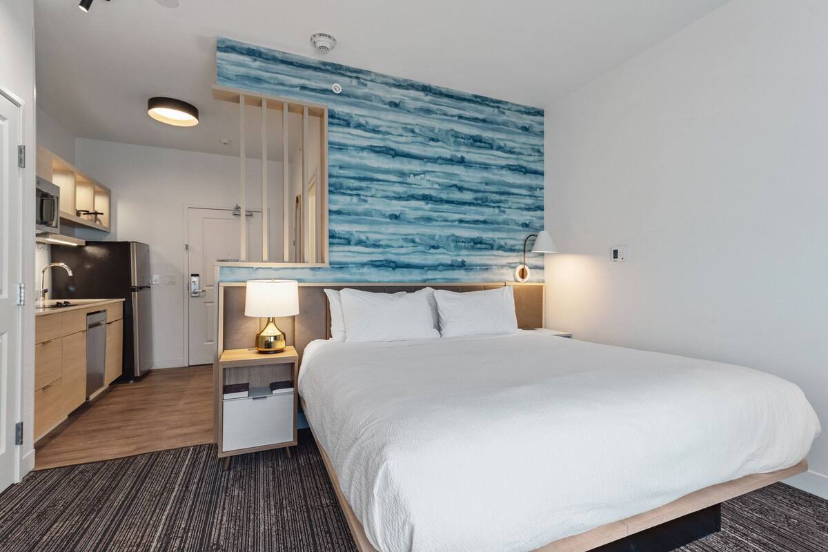 Una habitació d'hotel amb un llit, una taula de nit, una làmpada i vista a la cuina.