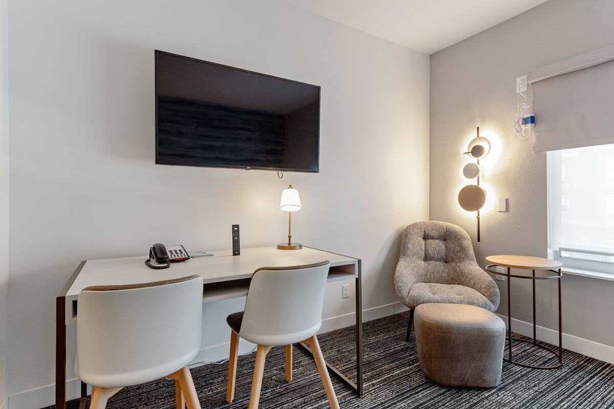 Habitació d'hotel moderna amb escriptori, dues cadires, un sofà, una taula petita i un televisor fixat a la paret.