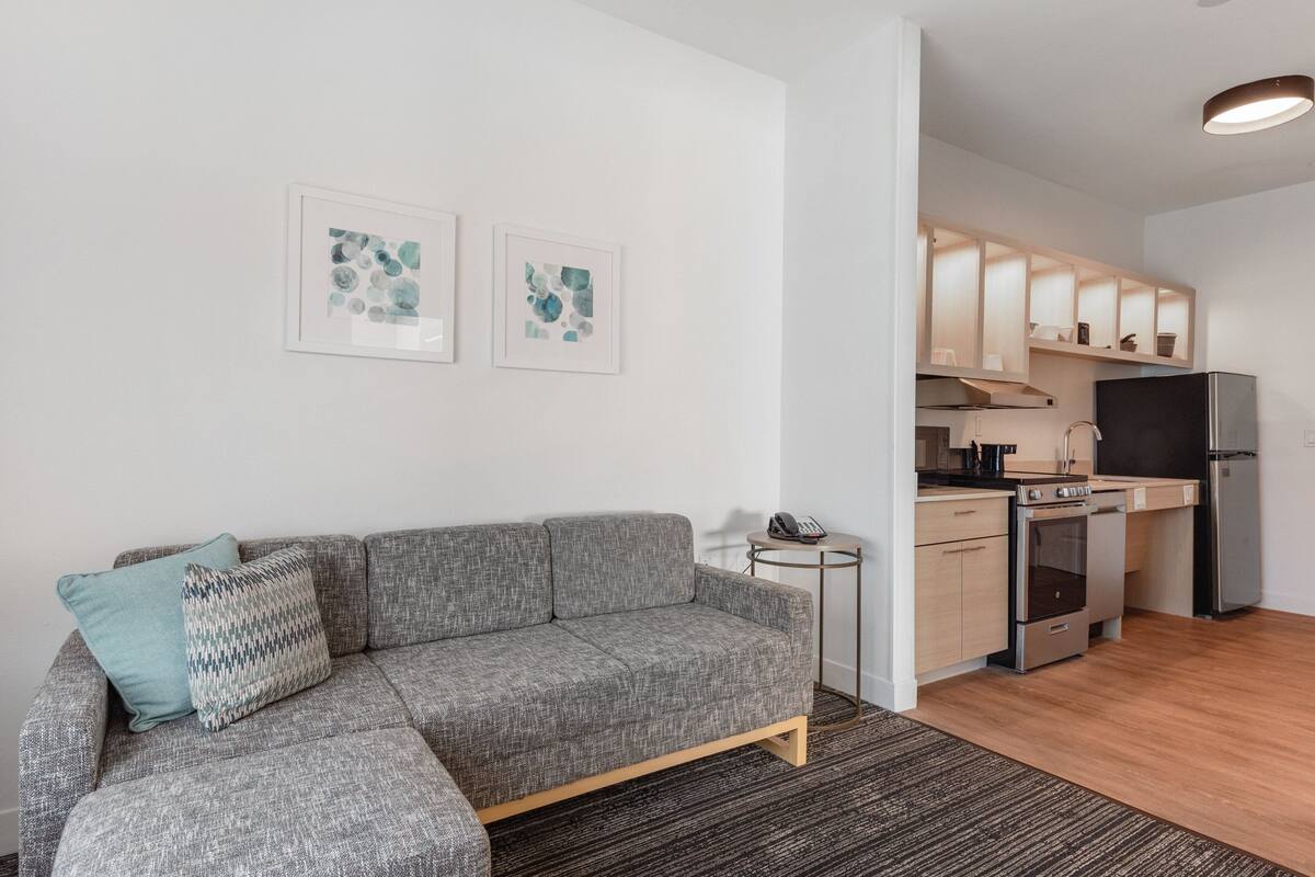 Un apartament modern amb un sofà gris, una taula auxiliar petita i una zona de cuina amb armaris de fusta i electrodomèstics.