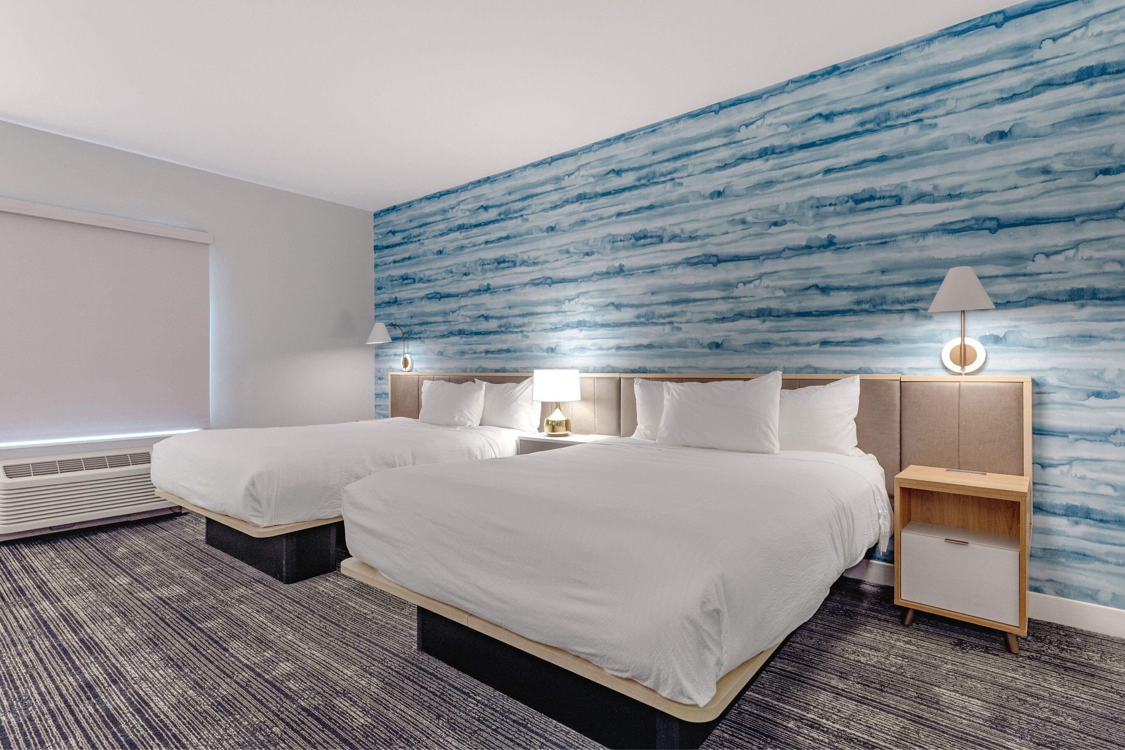 A hotel room with two beds, a large window, a wall with a blue abstract design, and a nightstand with a lamp.