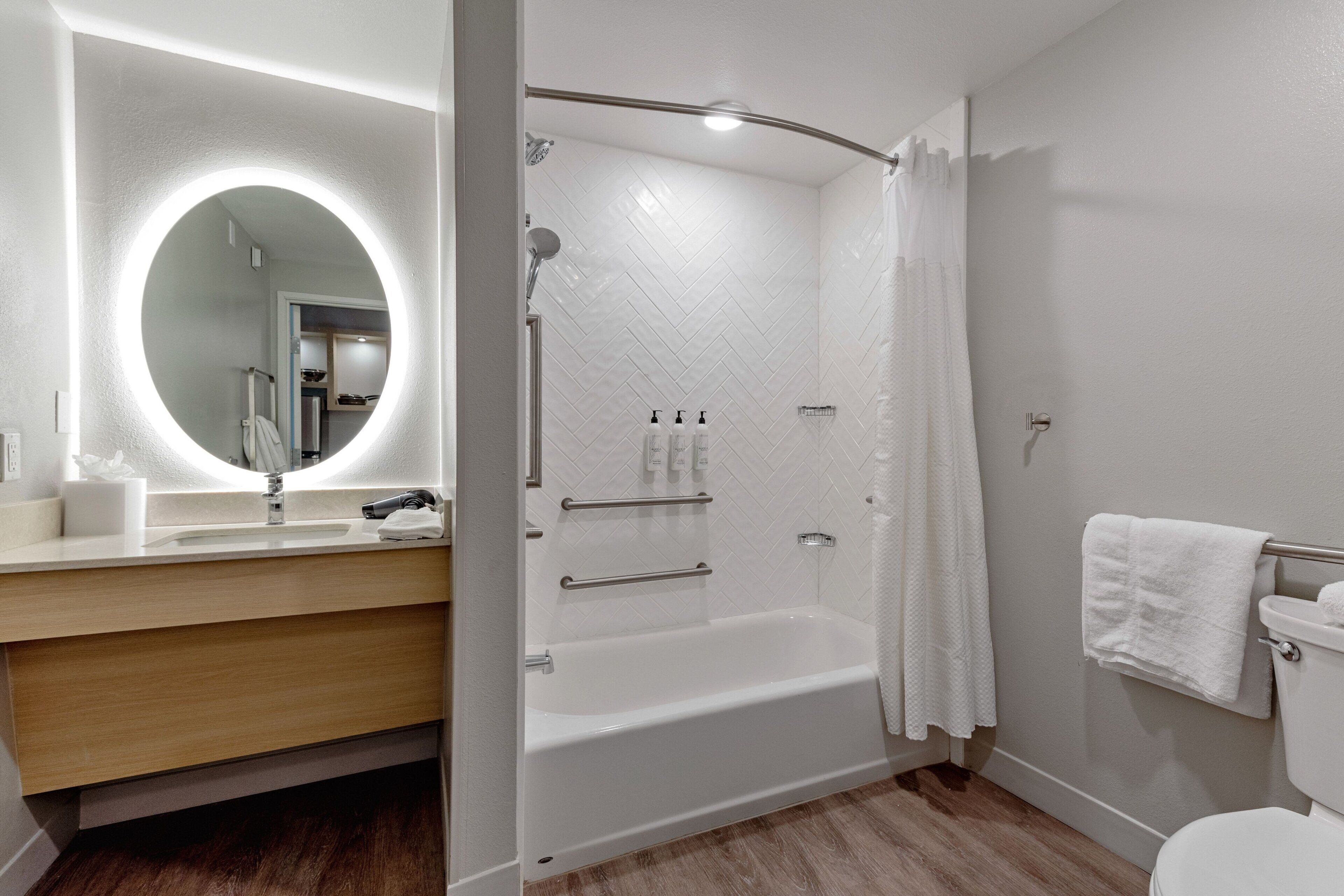 A modern bathroom with a white bathtub, a round mirror with a light, a wooden vanity, and a toilet with a towel hanging on the wall.