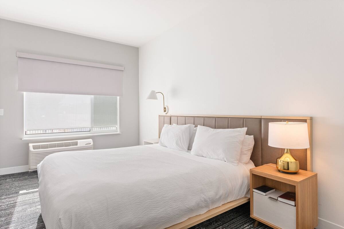 Habitació d'hotel amb un llit gran, una tauleta de nit amb una làmpada, una finestra amb estoreta i un radiador.