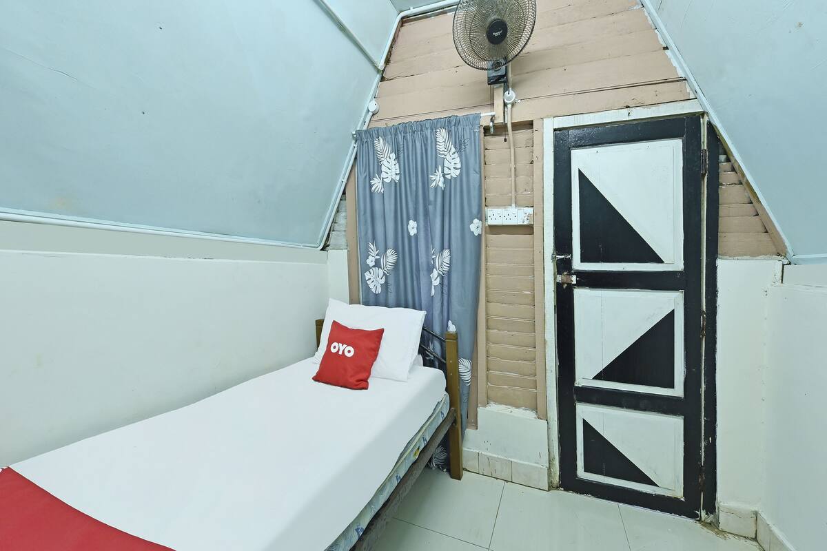 Una habitación pequeña con cama individual, cama blanca, almohada roja, ventilador y puerta con diseños geométricos.