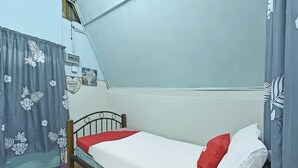Una habitación pequeña con cama individual, estructura metálica, cortina floral gris y colcha blanca con almohada roja.