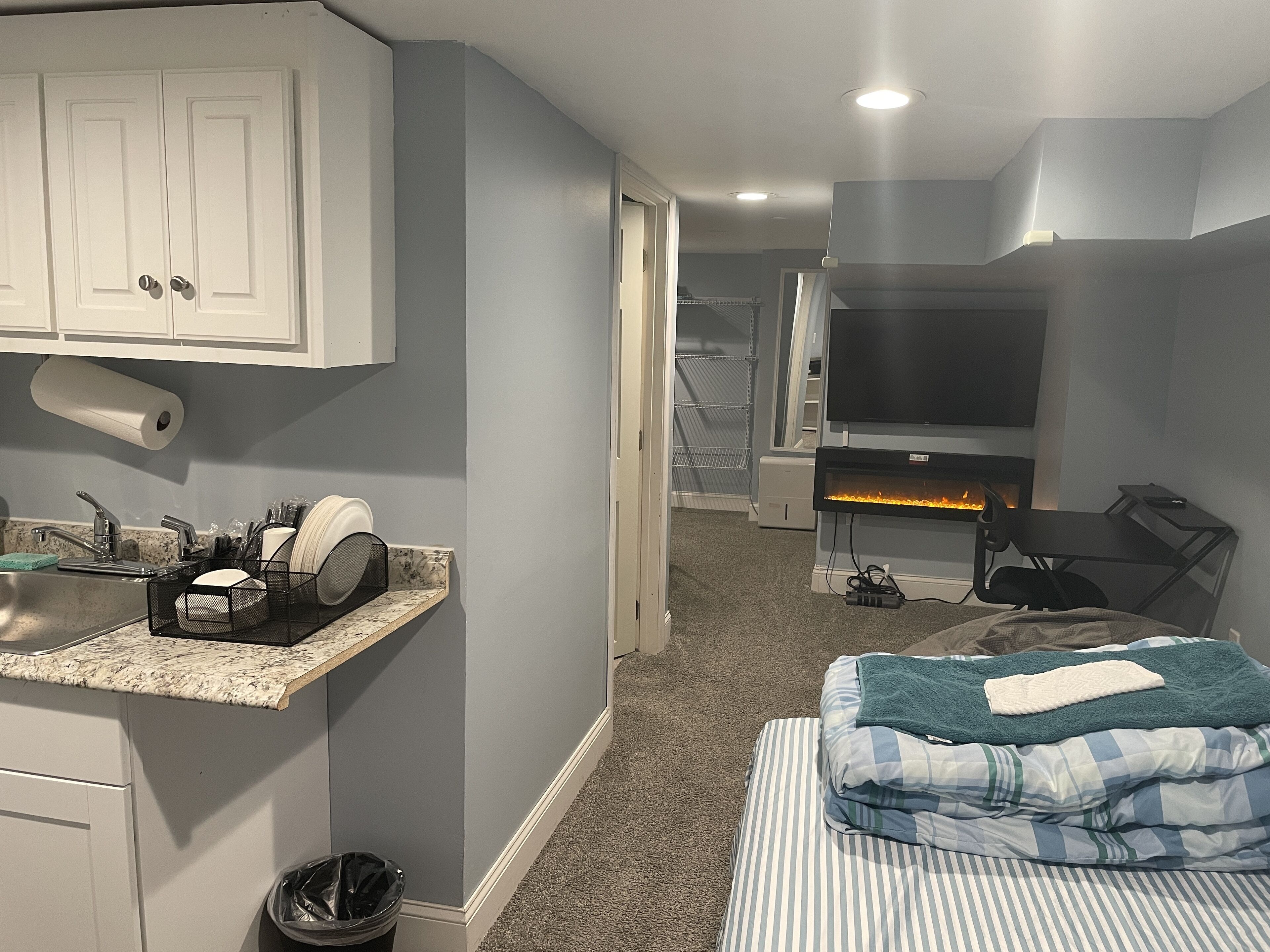 A compact hotel room with a kitchenette, a bed with striped bedding, a TV, and a fireplace.