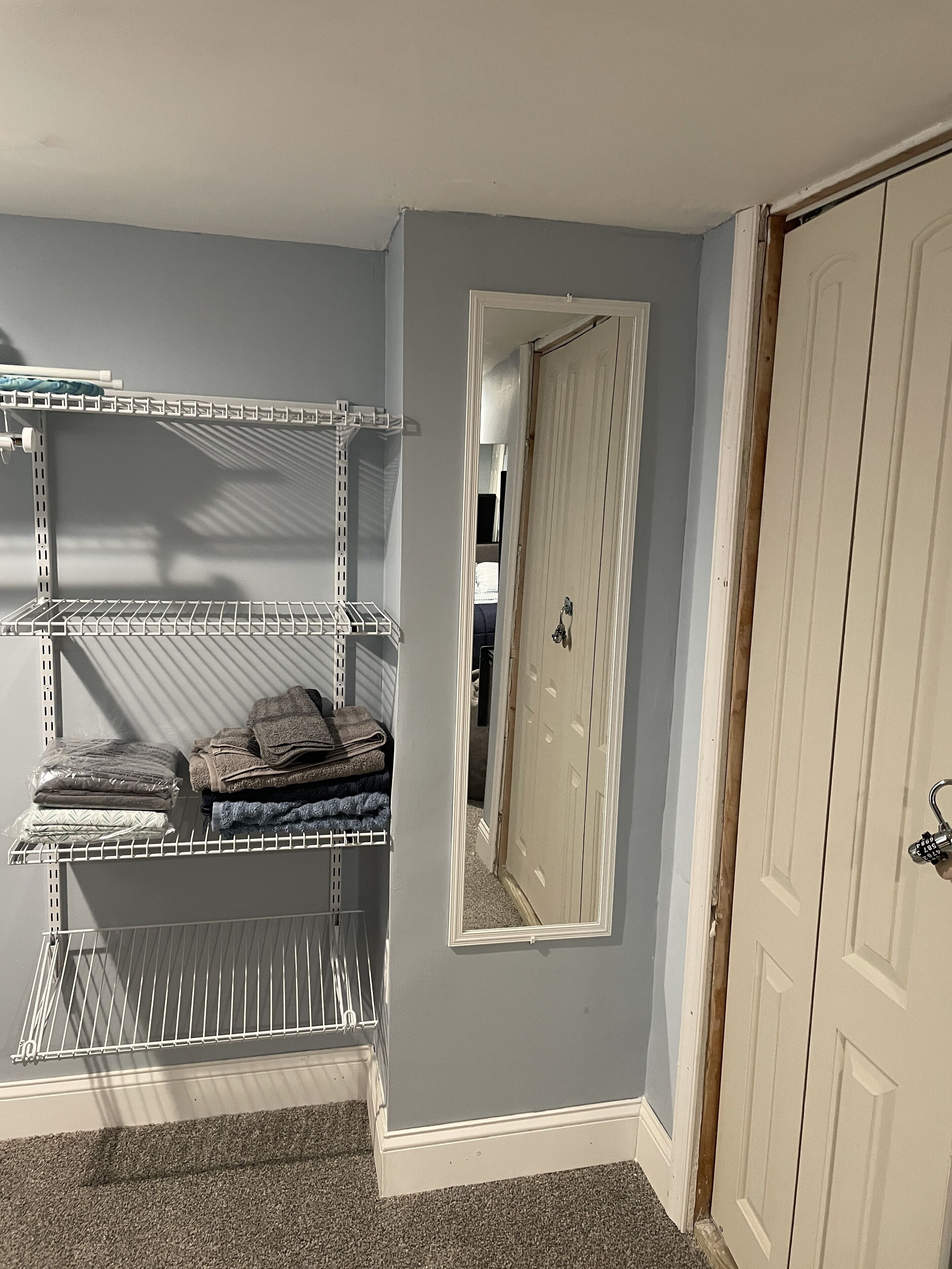 Una habitación con alfombra gris, una puerta blanca con cerradura y un espejo en la pared.