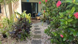 Caminho no jardim que leva à entrada de um edifício, cercado por plantas variadas e uma área de estar.