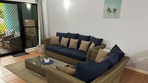 Uma sala de estar com um sofá de vime, uma mesa de centro de vidro e uma pequena planta em vaso.