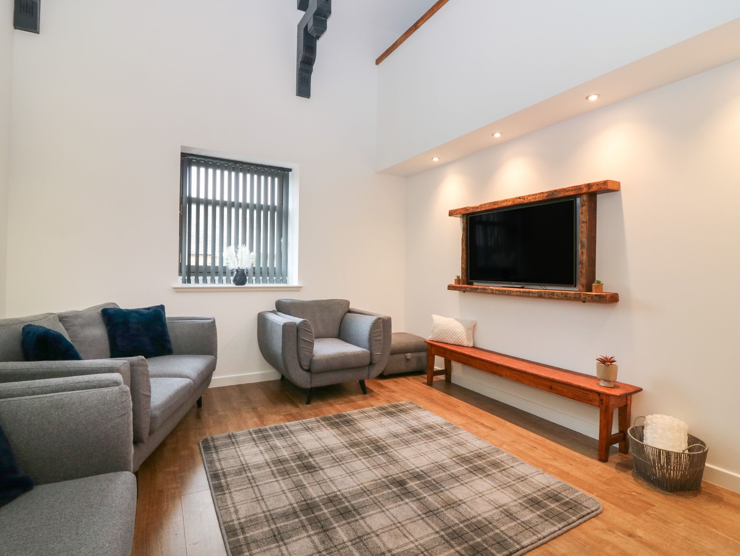 Una sala de estar moderna con un sofá gris, un sillón gris más pequeño, un tapete escocés, un banco de madera y un televisor montado en la pared.
