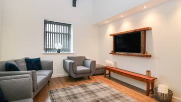 Una sala de estar moderna con un sofá gris, un sillón gris más pequeño, un tapete escocés, un banco de madera y un televisor montado en la pared.