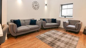 A living room with a grey sofa set, a plaid rug, a wall clock, and a window with bars. - The Reception Room at The Institute Executive Apartments (Keith)