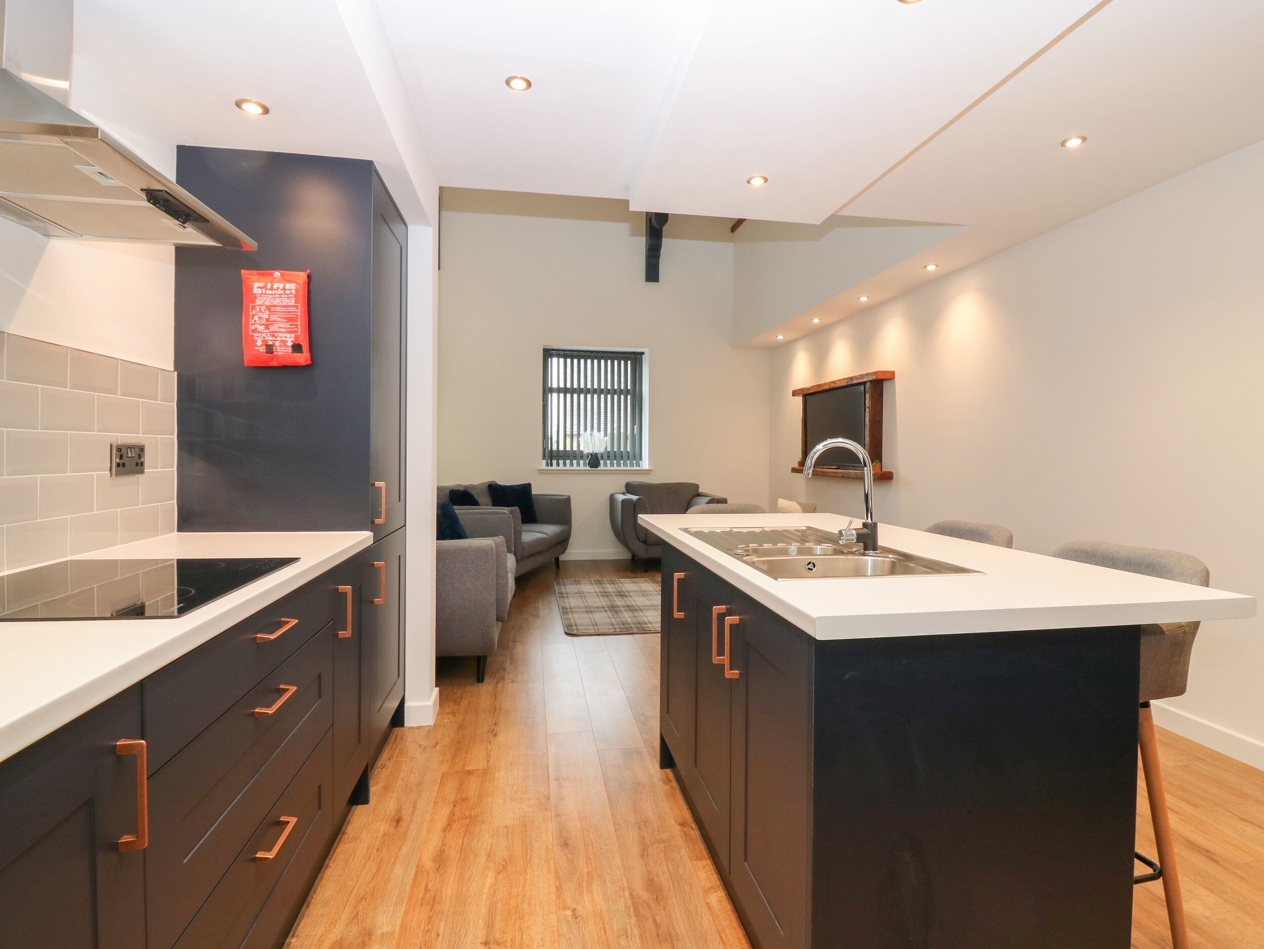 Una cocina moderna con armarios oscuros, encimeras blancas y una isla central con un lavabo. Se ve una ventana con persianas y un sofá al fondo.