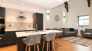 A modern kitchen with a central island, bar stools, and a dining table. There is a living area with sofas and a large window. - The Reception Room at The Institute Executive Apartments (Keith)