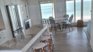 A modern kitchen with a marble countertop, white cabinetry, and a stainless steel refrigerator. There is a dining area with wicker chairs and a table, and a large window offering a view of the ocean.