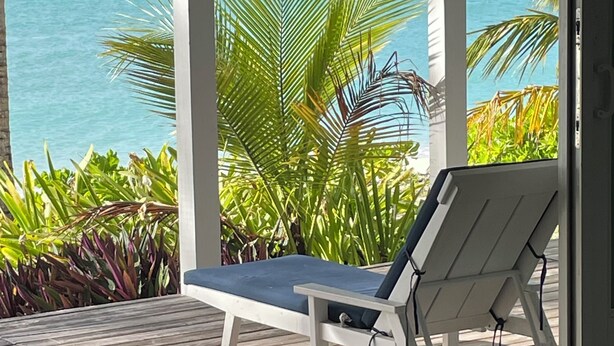 A wooden deck with a white lounge chair and a dark sofa, overlooking a turquoise sea with palm trees.