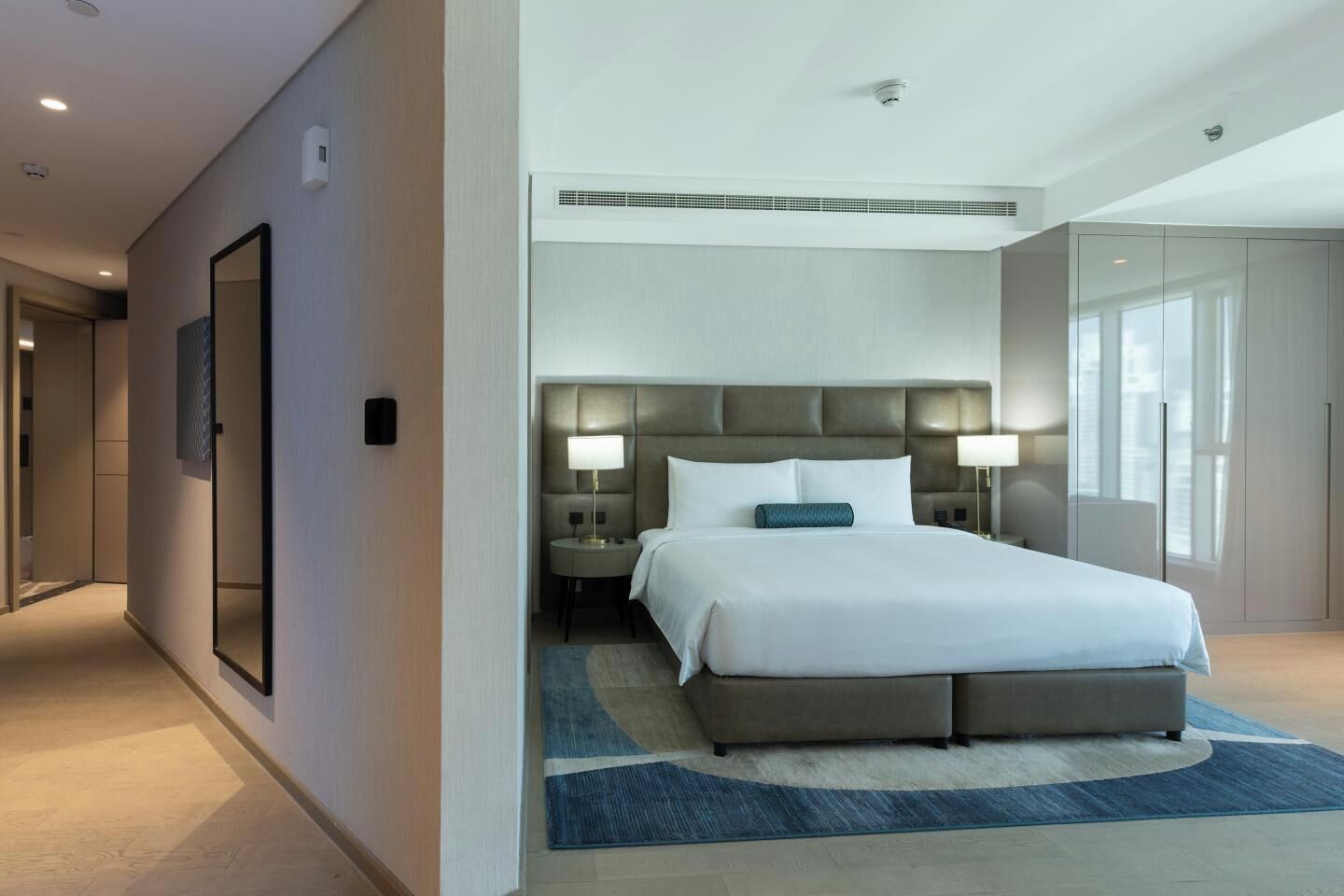 Quarto de hotel moderno com uma cama grande, mesas de cabeceira, um espelho e vista da cidade.