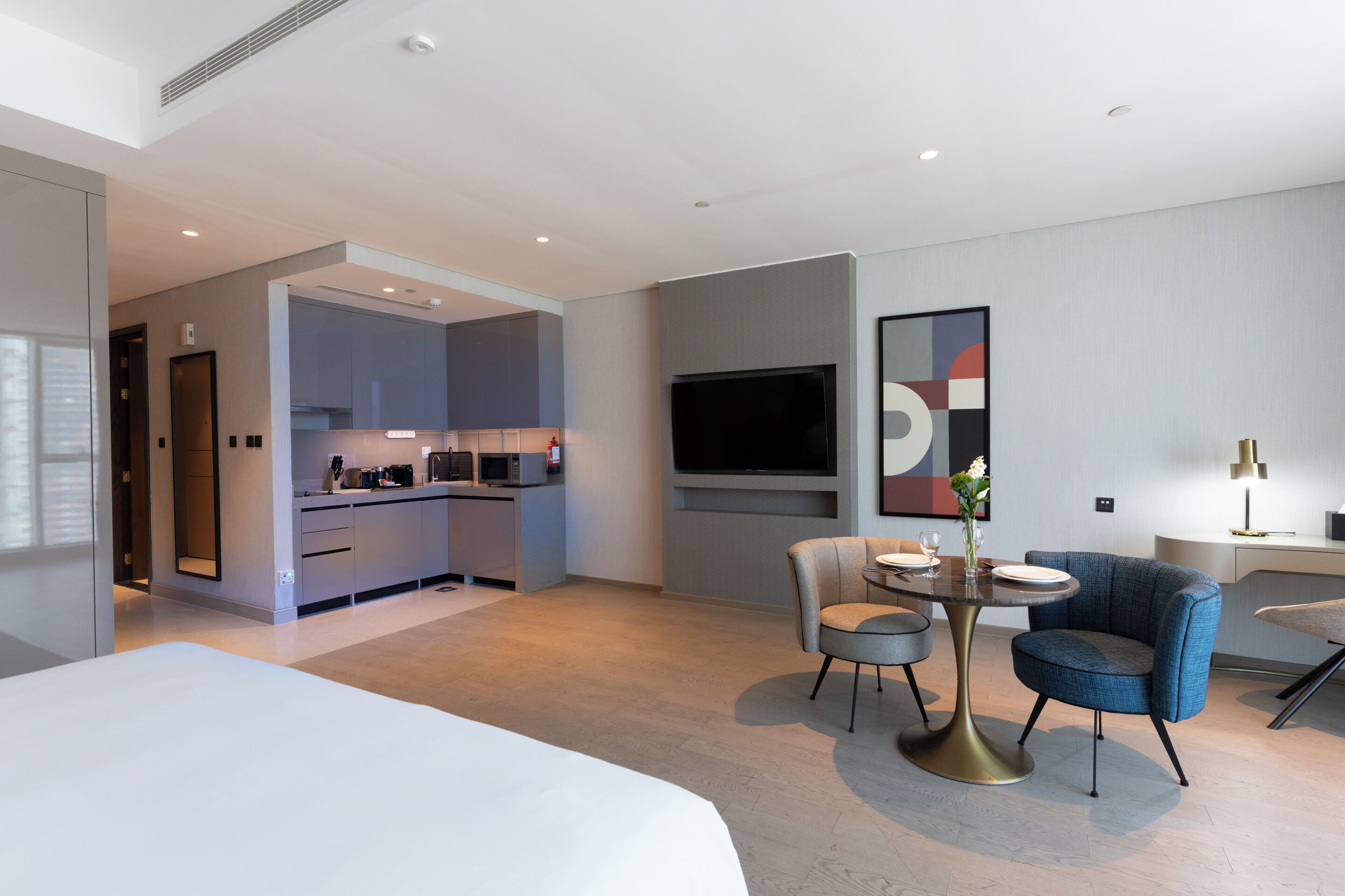 Een moderne hotelkamer met een groot bed, een zithoek met twee stoelen, een tv aan de wand en een kitchenette op de achtergrond.