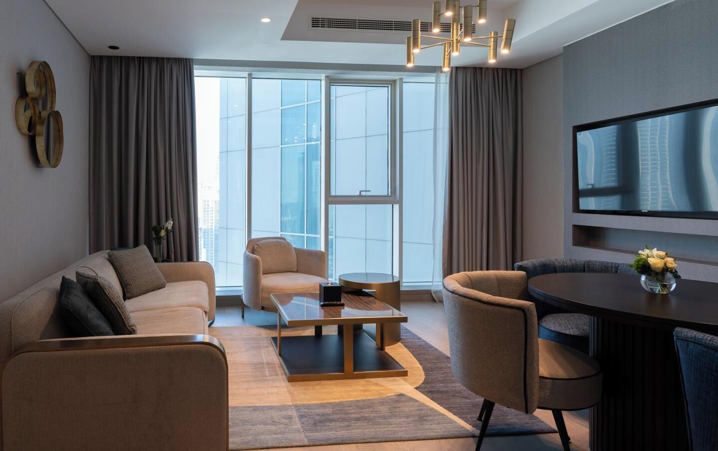 Una habitación de hotel moderna con sofá, sillón, mesa de comedor y televisor en la pared.