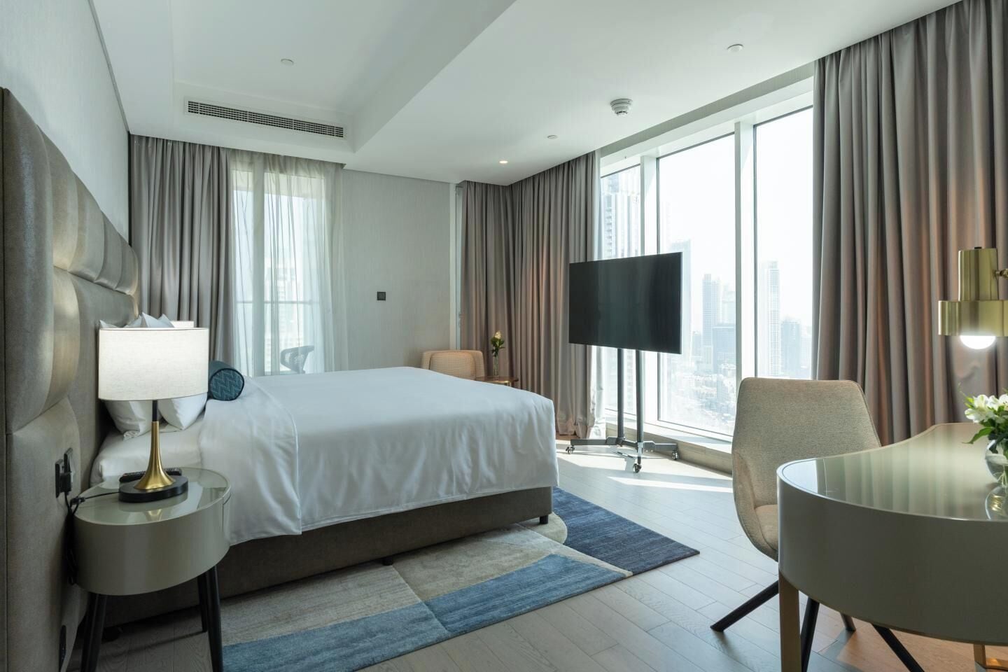 A hotel room with a bed, a desk, a chair, a TV, and large windows.
