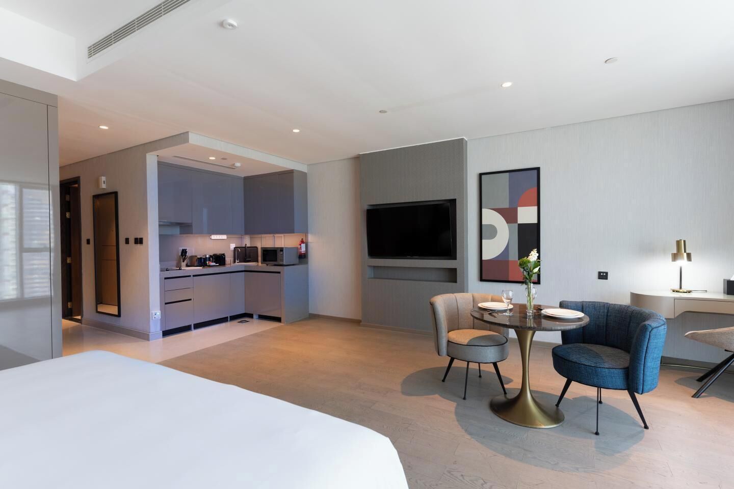 A modern hotel room with a large bed, a dining area with a round table, a TV mounted on the wall, and a kitchenette.
