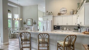Una cocina con armarios blancos, una isla central con taburetes y un refrigerador, además de un reloj en la pared.
