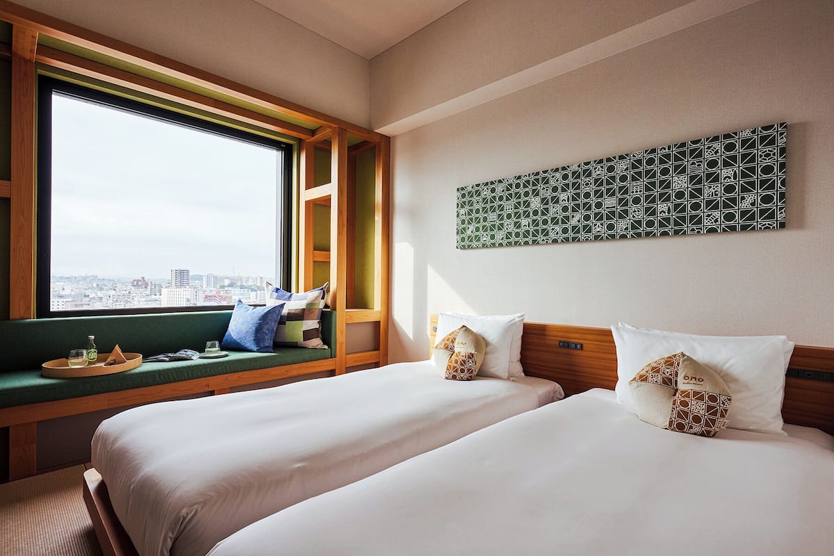 Une chambre d’hôtel avec deux lits, une fenêtre donnant sur la ville, un mur végétal et une œuvre d’art aux motifs géométriques.