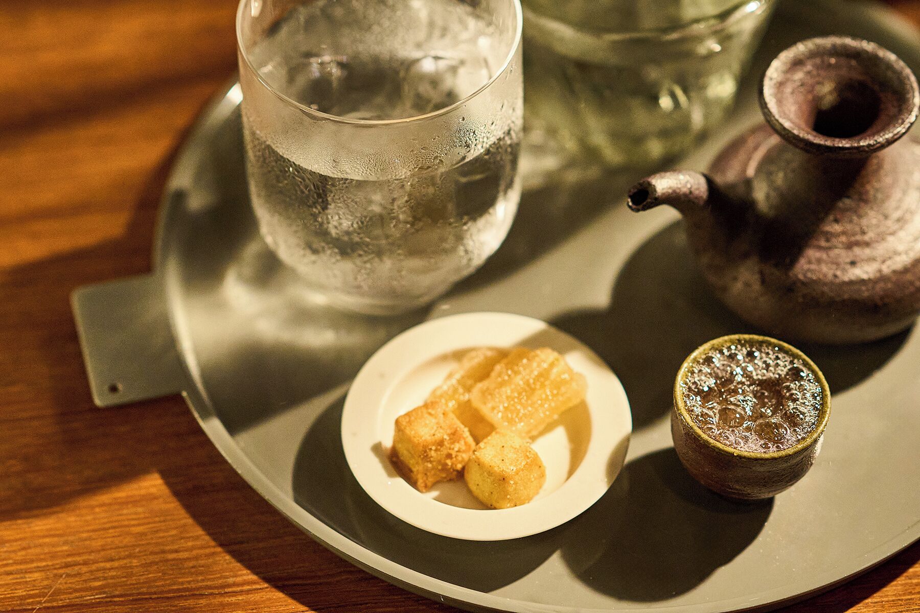 Una bandeja con un vaso de agua, un plato pequeño con dos cubitos de hielo y una taza con una bebida espumosa.