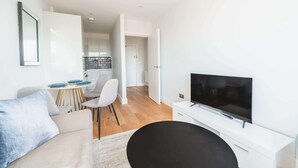 Ett modernt vardagsrum med en platt-TV, en soffa, en matplats med bord och stolar samt ett kök i bakgrunden.