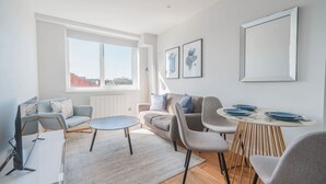 Ett modernt vardagsrum med en soffa, fåtöljer, ett soffbord och ett matbord med plats för fyra personer. Ett stort fönster släpper in naturligt ljus och ger utsikt över byggnaderna utanför.
