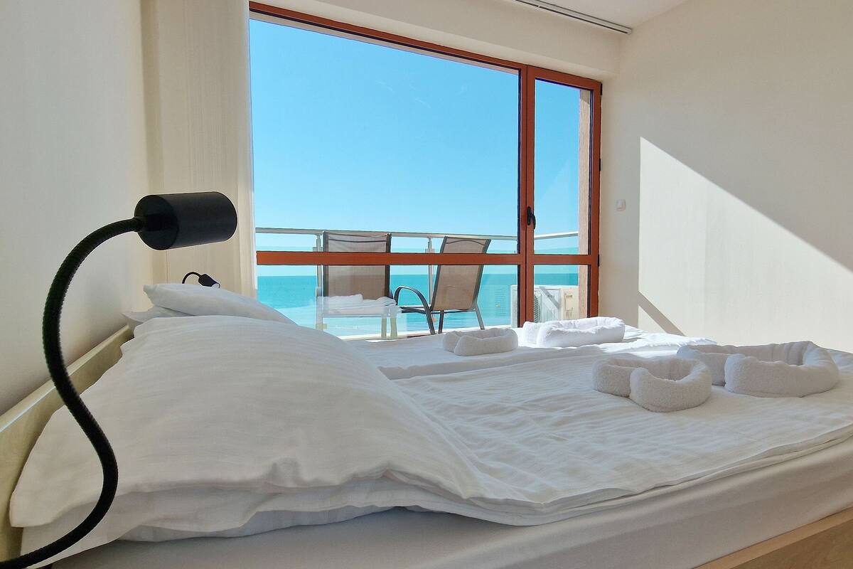 A hotel room with a bed, a desk, and a view of the ocean through large windows.