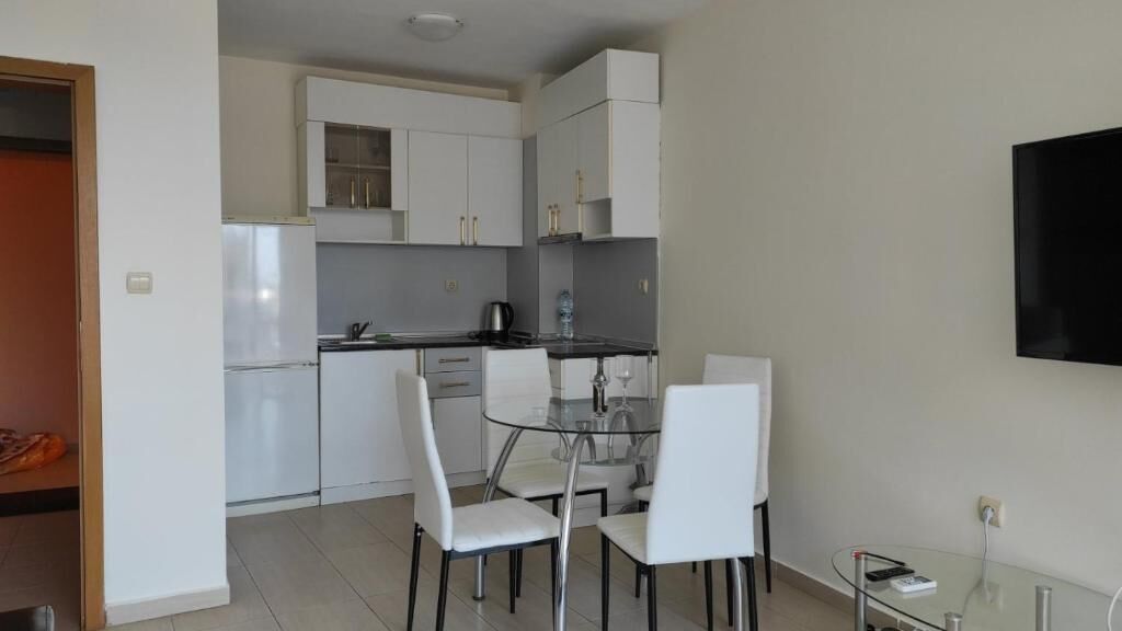 A compact kitchen with white cabinets, a glass dining table, and a TV mounted on the wall.