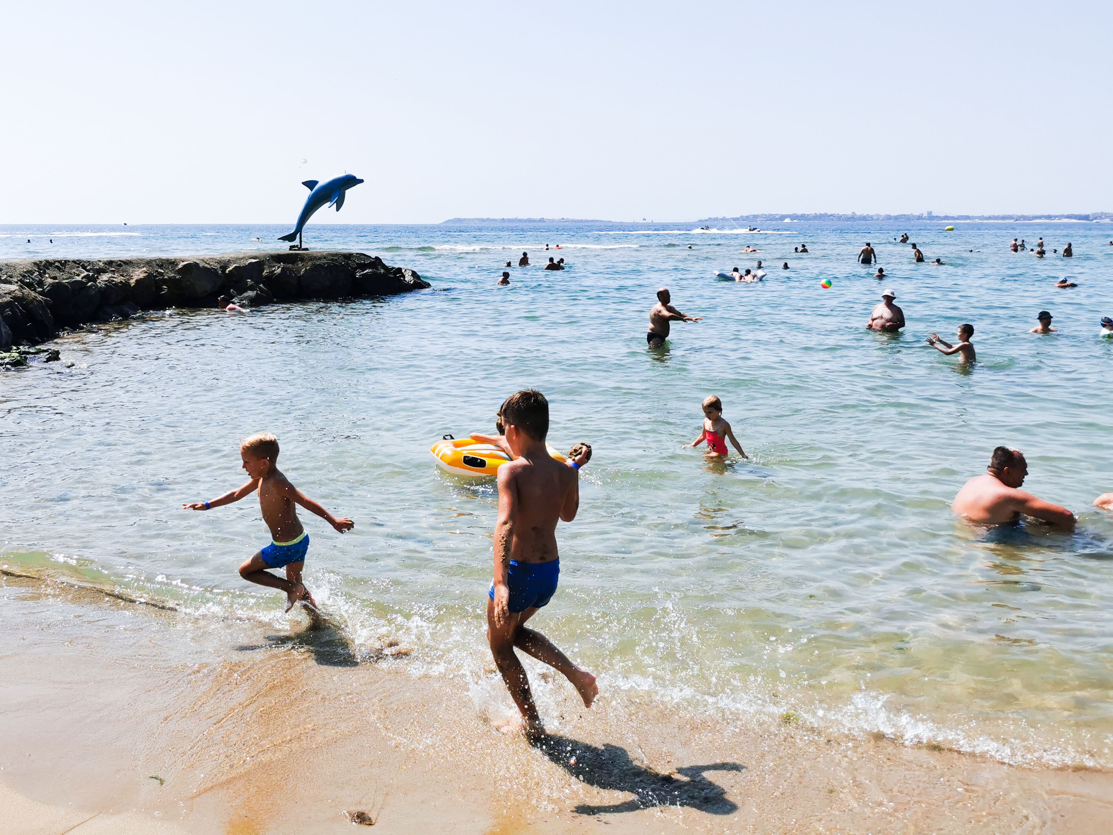 A beach scene with people swimming and playing in the water, a dolphin statue, and a rocky shoreline.