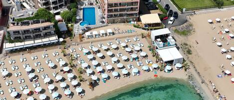 Vista aérea de una playa con numerosas tumbonas y sombrillas, una piscina y un edificio con varios balcones.