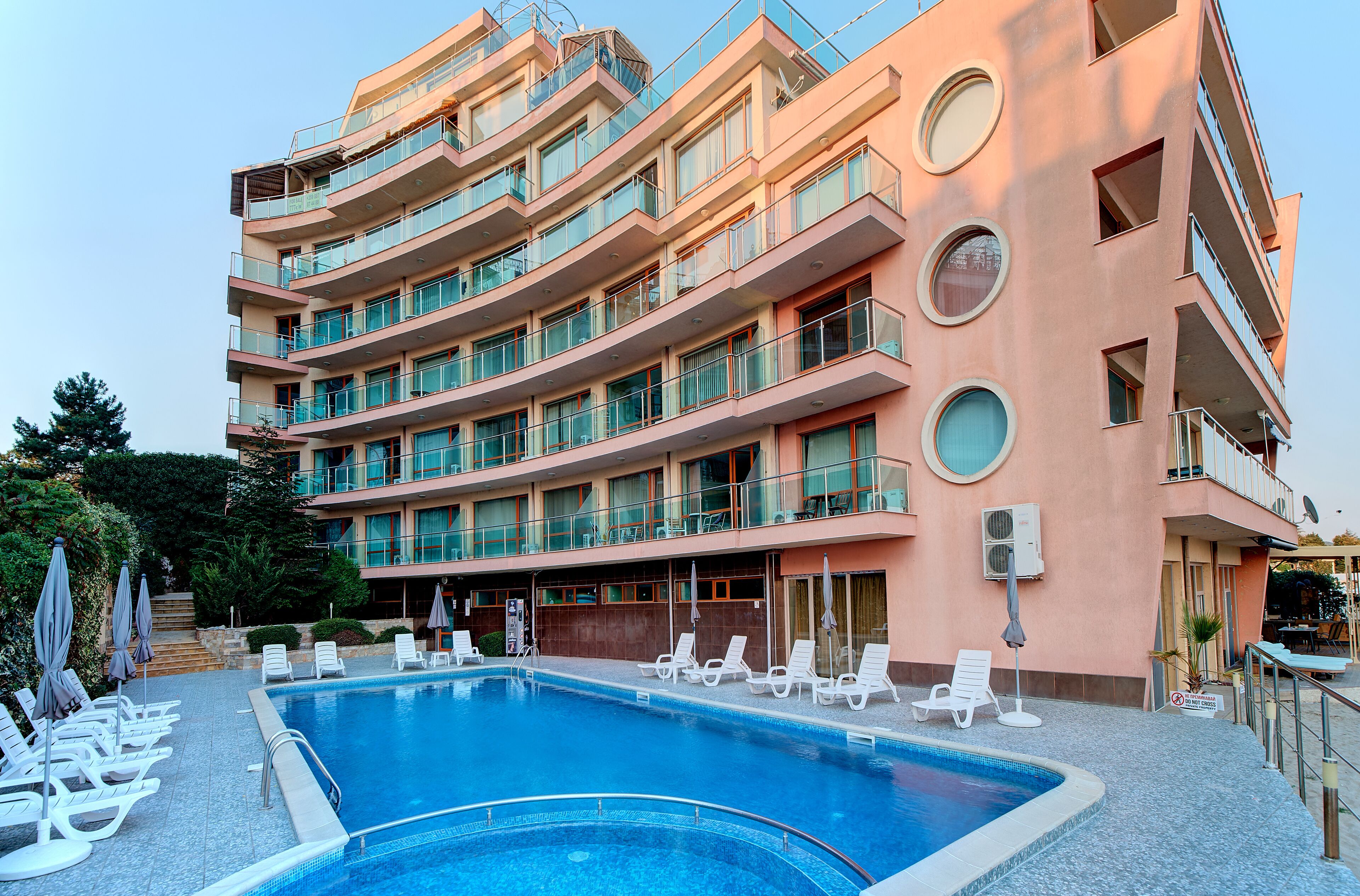 A multi-story hotel with a swimming pool, sun loungers, and a terrace with umbrellas.