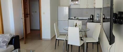 A compact living space with a kitchenette, a dining area with a glass table and chairs, a sofa, and a TV mounted on the wall.