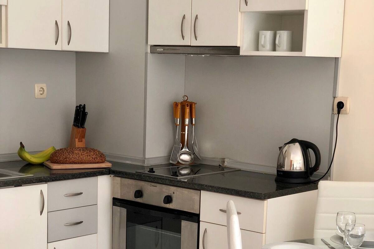 A modern kitchen with white cabinets, a stainless steel oven, a glass table with chairs, and a kettle on the counter.