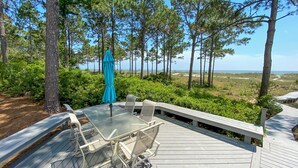 A wooden deck with a glass-top table and chairs, surrounded by greenery and overlooking a beach.