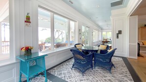 A bright, sunlit room with large windows, a glass-top table, and blue wicker chairs.