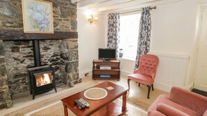 A cozy living room with a stone fireplace, a television, a plush armchair, and a coffee table. - Tudor Cottage (Conwy)